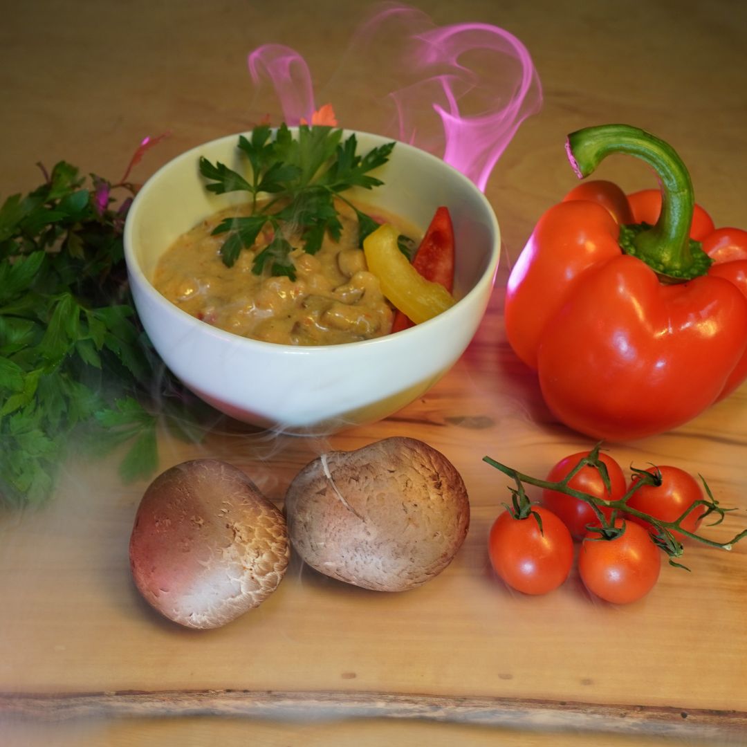 Schüssel cremige Pilzsuppe mit Petersilie und Paprikastreifen, umgeben von Champignons, Kirschtomaten, Petersilie und roter Paprika auf einem Holztisch; dekorativer rosa Dampf steigt auf.
