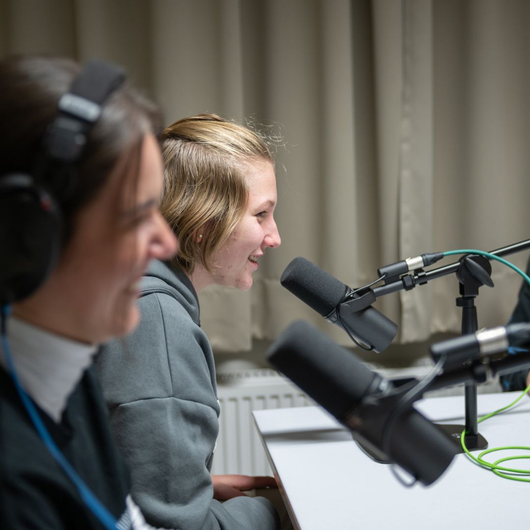 Zwei Personen nehmen in einem Podcast- oder Radiostudio ein Gespräch auf und sprechen lächelnd in Studiomikrofone.