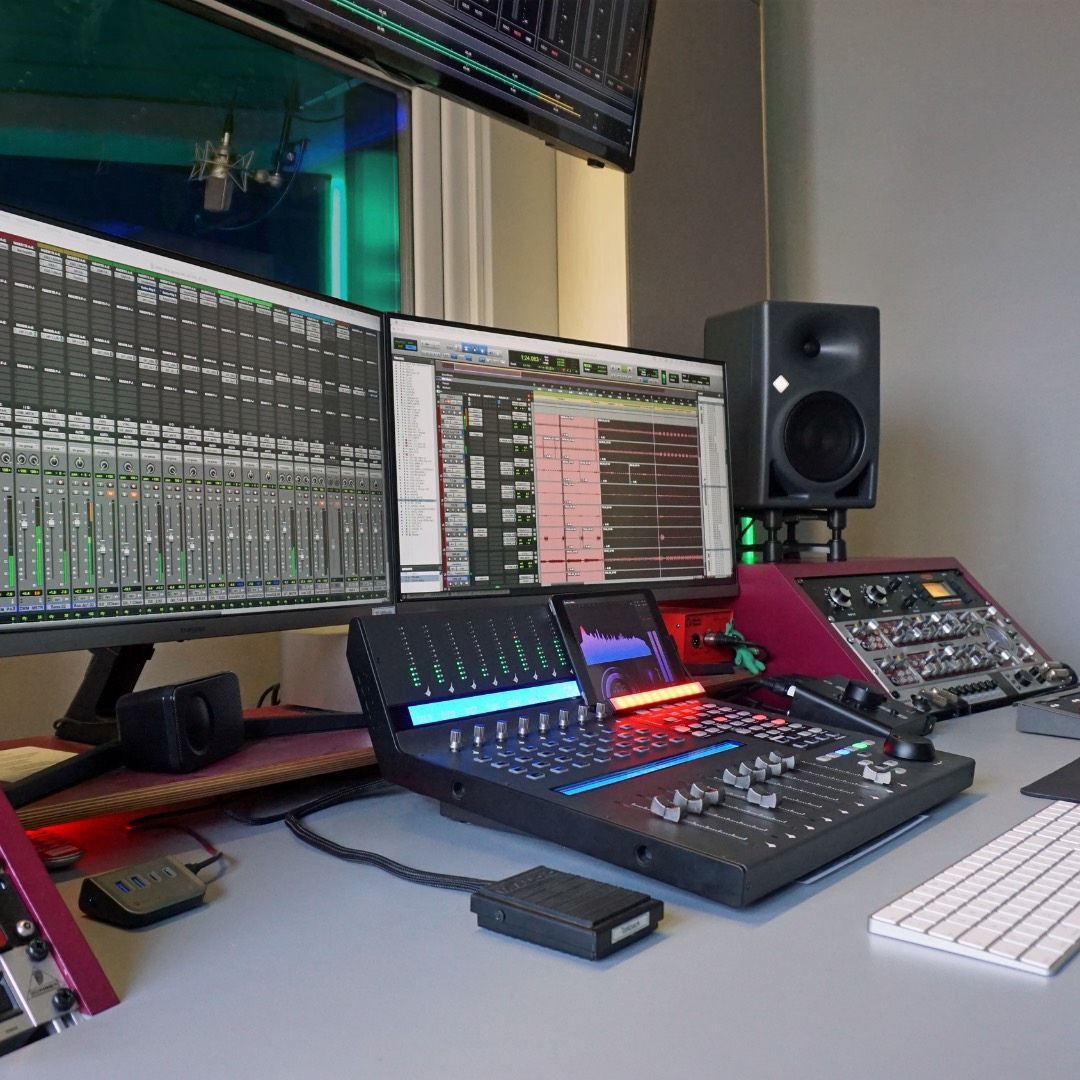 Regieplatz eines Tonstudios mit Mischpult, mehreren Monitoren mit Audio-Software, Studiolautsprecher und Blick in die Aufnahmekabine.