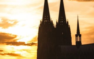 Der Kölner Dom als dunkle Silhouette mit zwei hohen Türmen vor einem warm leuchtenden Abendhimmel in Gelb- und Orangetönen.