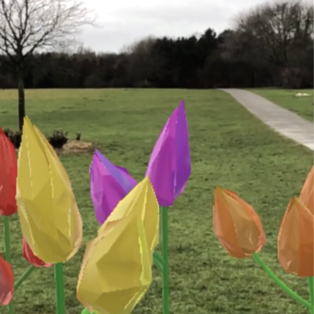 Bunte, geometrische Blumen stehen auf einer Wiese in einem Park, mit einem geschwungenen Weg, kahlen Bäumen und bewölktem Himmel im Hintergrund.