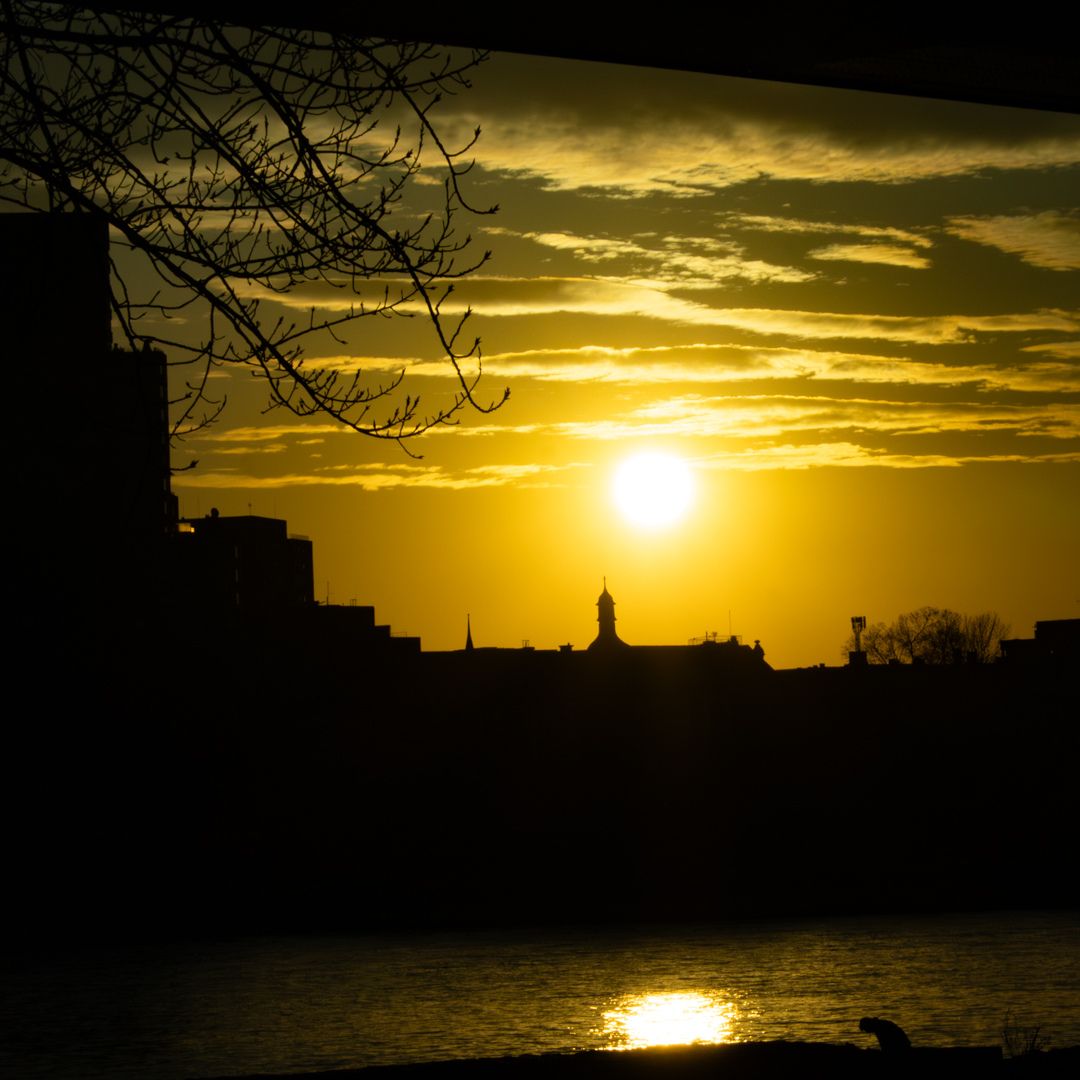 Goldener Sonnenuntergang über einer silhouettierten Stadt mit Turm, spiegelndem Fluss und einer sitzenden Person am Ufer.
