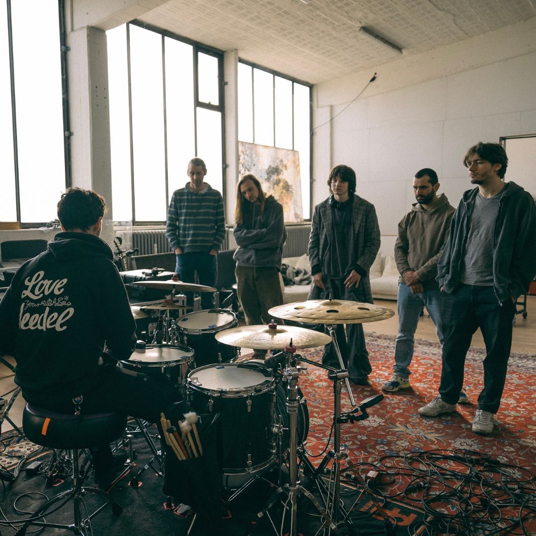 Ein Schlagzeuger spielt vor fünf Männern in einem hellen Proberaum mit großen Fenstern, Kabeln und Instrumenten auf einem roten Teppich.